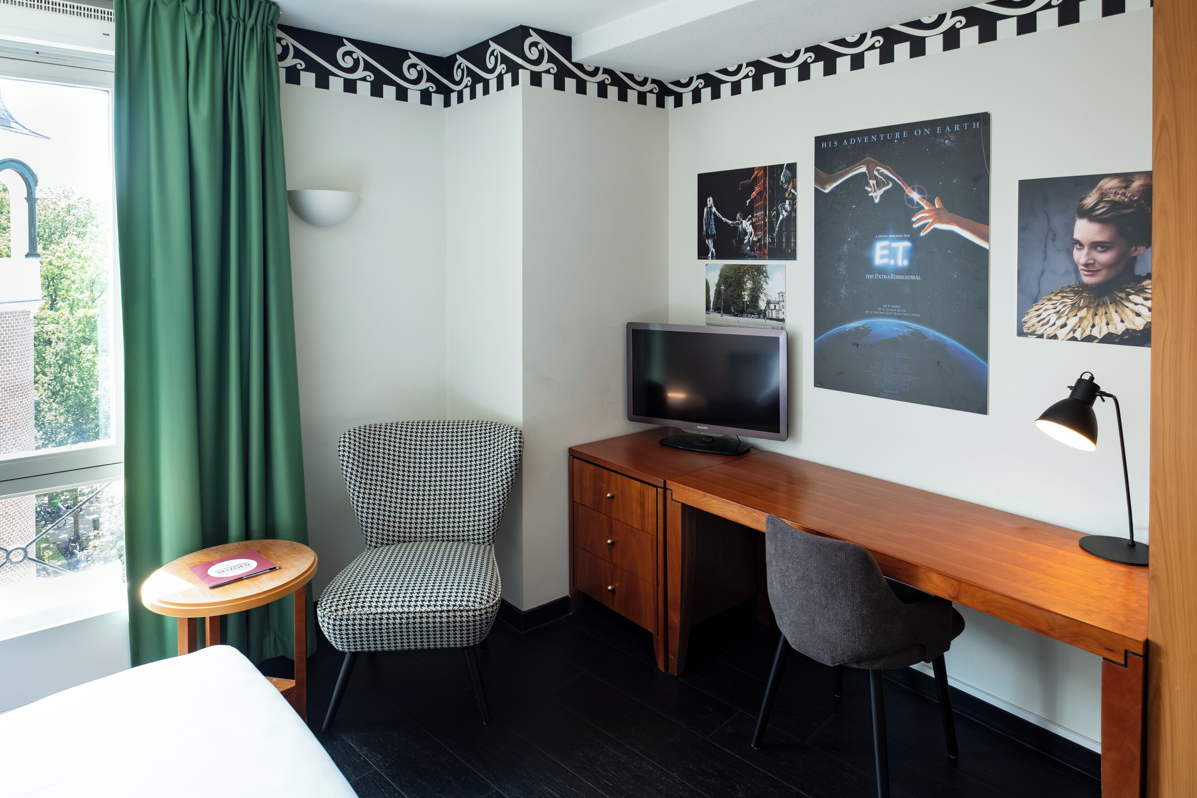 Habitación de hotel con cama, escritorio con computadora, silla, mesita y ventana con cortinas.