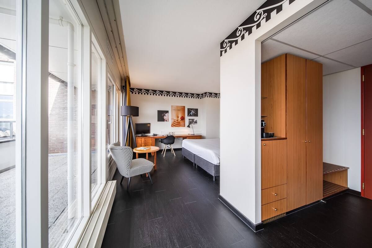 A modern hotel room with a large window, a desk with a computer, a bed, a wardrobe, and a small seating area.
