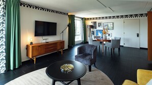 A modern living room with a flat-screen TV, a wooden cabinet, a round coffee table, and a dining area with chairs and a table. - Hotel Theater Figi (Zeist)