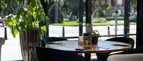 A dining area with a table set for two, a potted plant, and a view of the street outside.