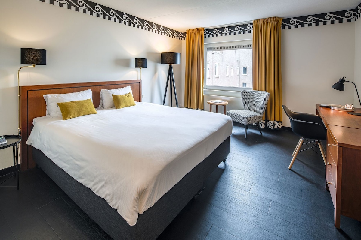 A hotel room with a large bed, two bedside lamps, a desk, a chair, and a view of buildings through the window.