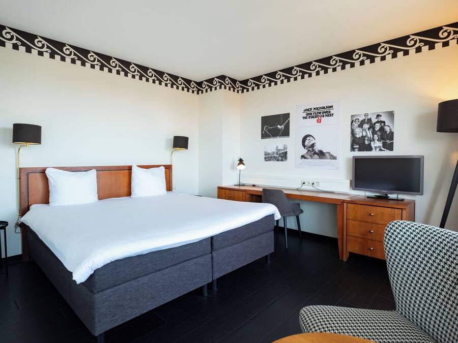 Una habitación de hotel con cama, escritorio, silla, televisor y decoración en la pared.