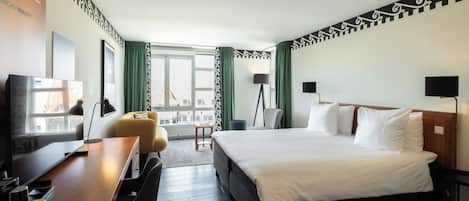 Habitación de hotel con cama, escritorio, silla, lámpara y vista del paisaje urbano a través del ventanal.