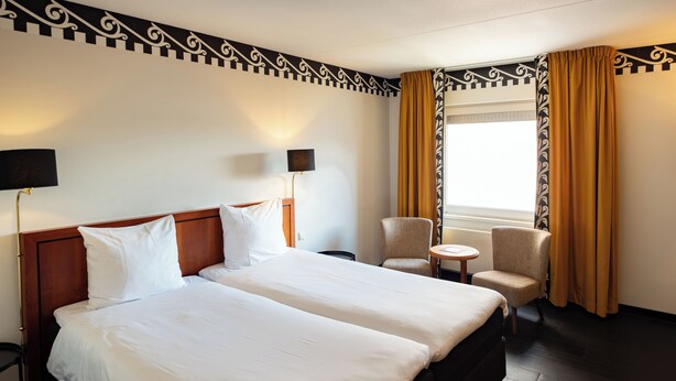 A hotel room with two beds, a small round table, a chair, and a window with curtains. - Hotel Theater Figi (Zeist)