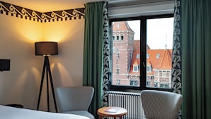 A hotel room with a bed, two houndstooth patterned armchairs, a small round table, a floor lamp, and a window with a view of a red-roofed building. - Hotel Theater Figi (Zeist)