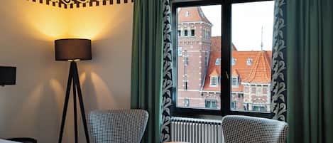 A hotel room with a bed, two houndstooth patterned armchairs, a small round table, a floor lamp, and a window with a view of a red-roofed building.