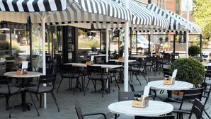 An outdoor seating area with round tables and chairs, covered by striped awnings, and a brick building in the background. - Hotel Theater Figi (Zeist)