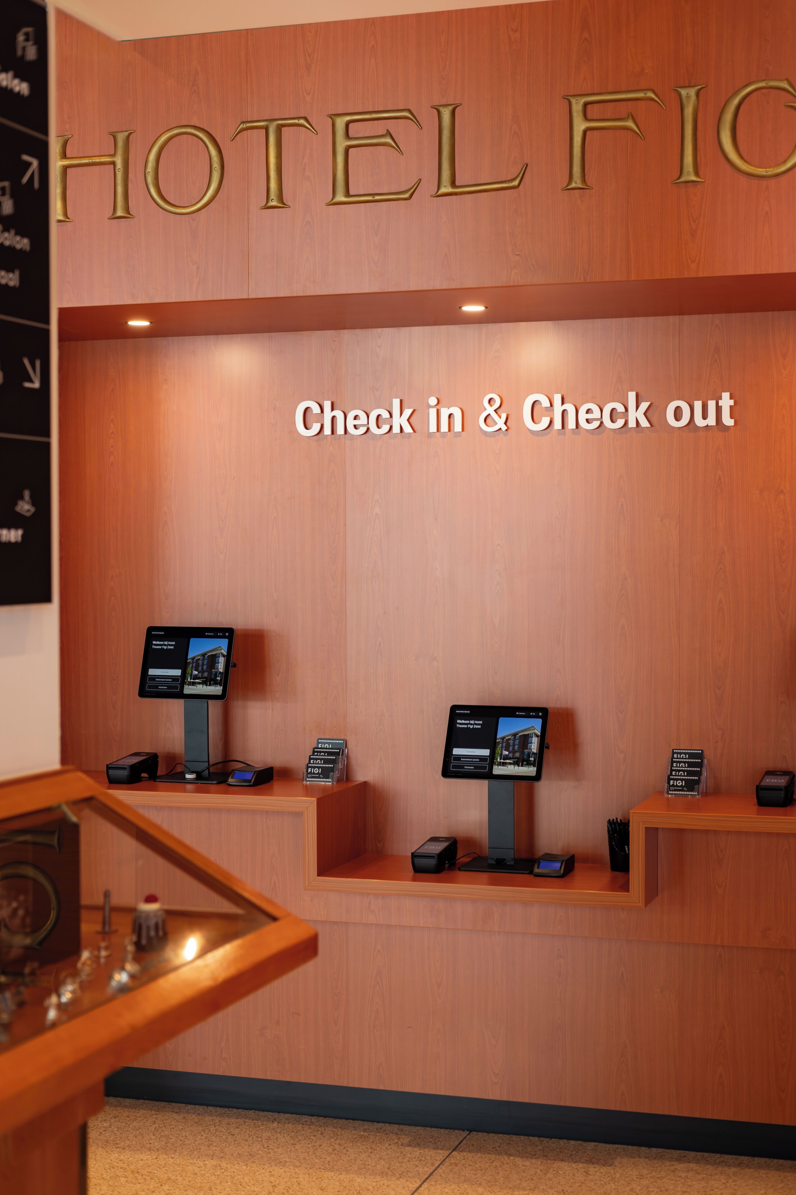 Zona de recepción de un hotel con un mostrador de madera, dos quioscos de check-in y un cartel que dice 'HOTEL FIC'.