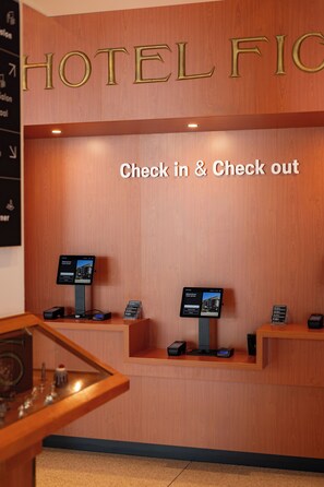 Een hotel receptie met een houten balie, twee incheckautomaten, en een bord met de tekst 'HOTEL FIC'.