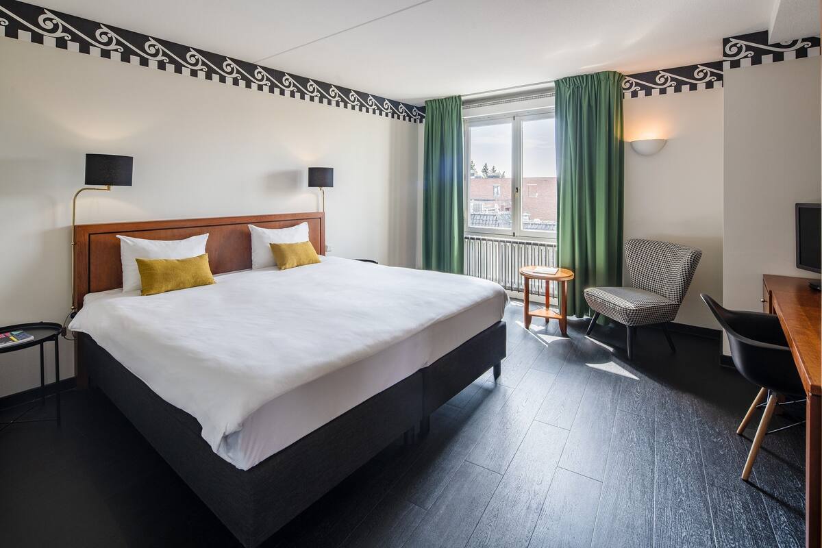 A hotel room with a large bed, two bedside tables, a chair, a desk, and a window with curtains.