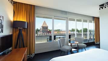 Quarto de hotel com cama, escrivaninha, cadeira, mesa e uma grande janela com vista para um edifício com torre.