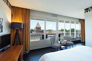 A hotel room with a bed, a desk, a chair, a table, and a large window offering a view of a building with a tower. - Hotel Theater Figi (Zeist)