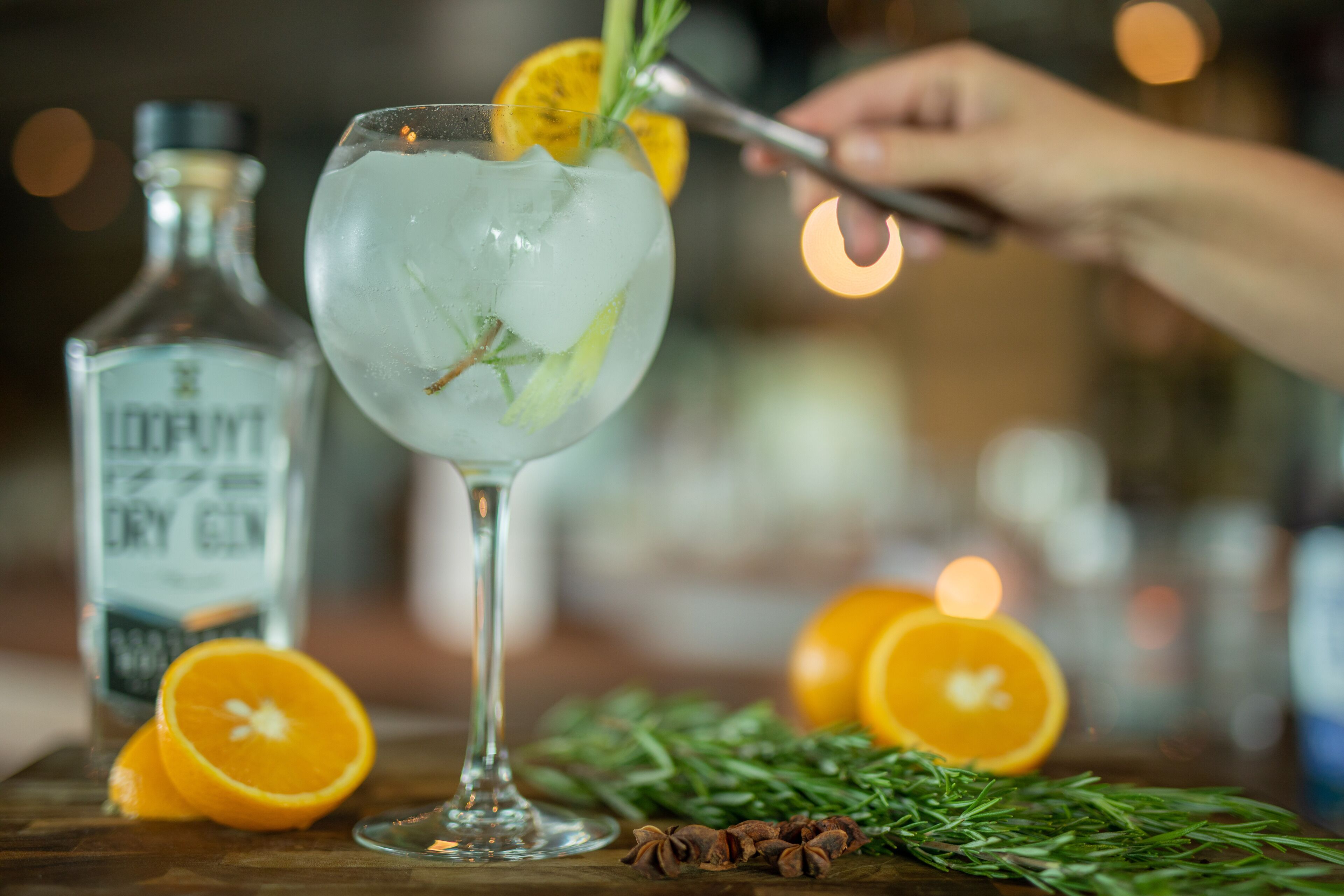 A cocktail glass with ice and a slice of orange, garnished with a sprig of rosemary, on a wooden surface.