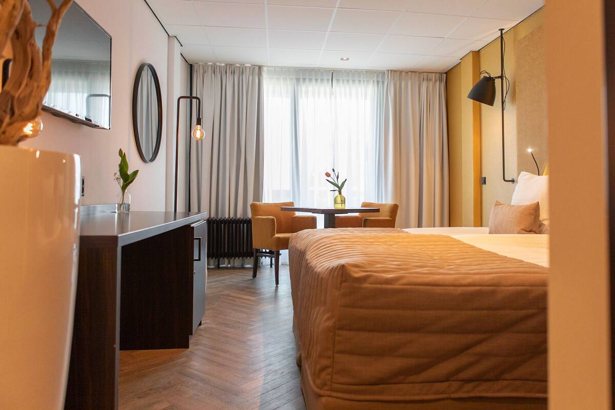 A hotel room with a bed, a desk, a chair, a table with a vase, and a window with curtains.