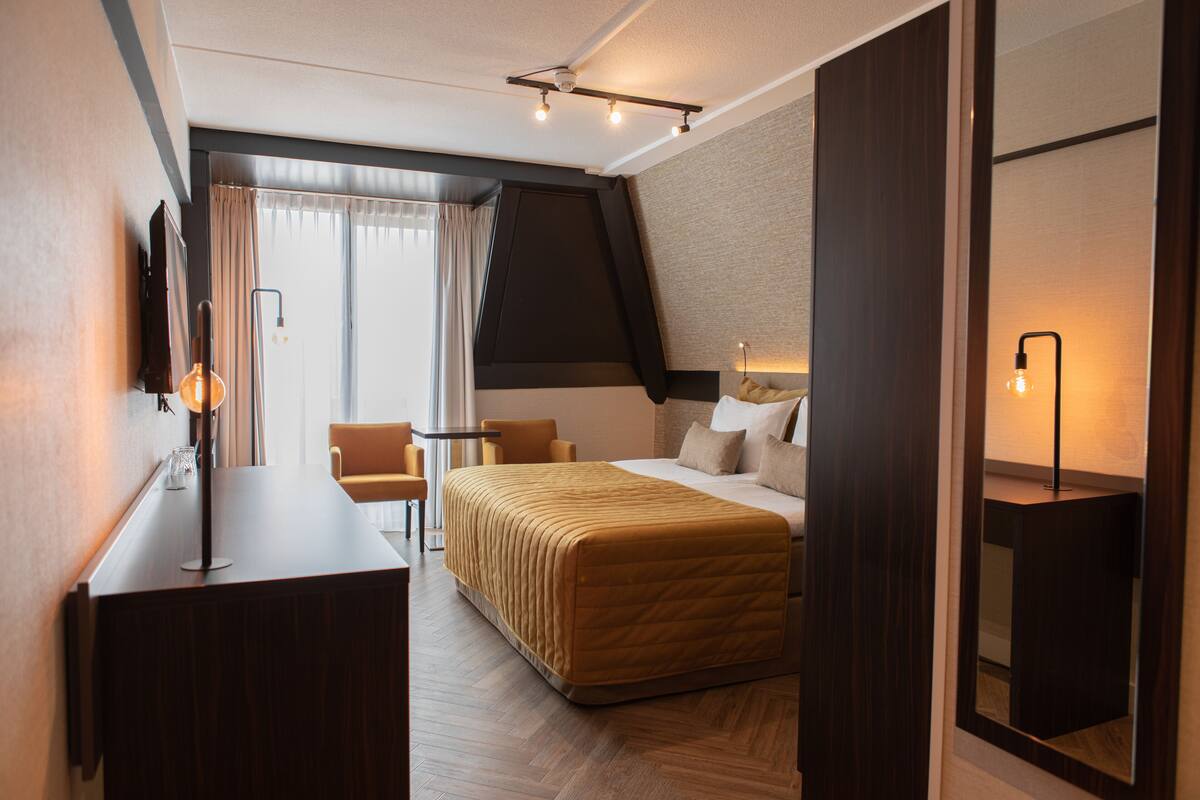 A hotel room with a bed, a desk, a chair, a lamp, and a large mirror.