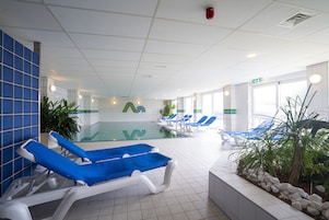 Un área de piscina con sillones de descanso azules y blancos, piso de baldosas y una pared de vidrio.