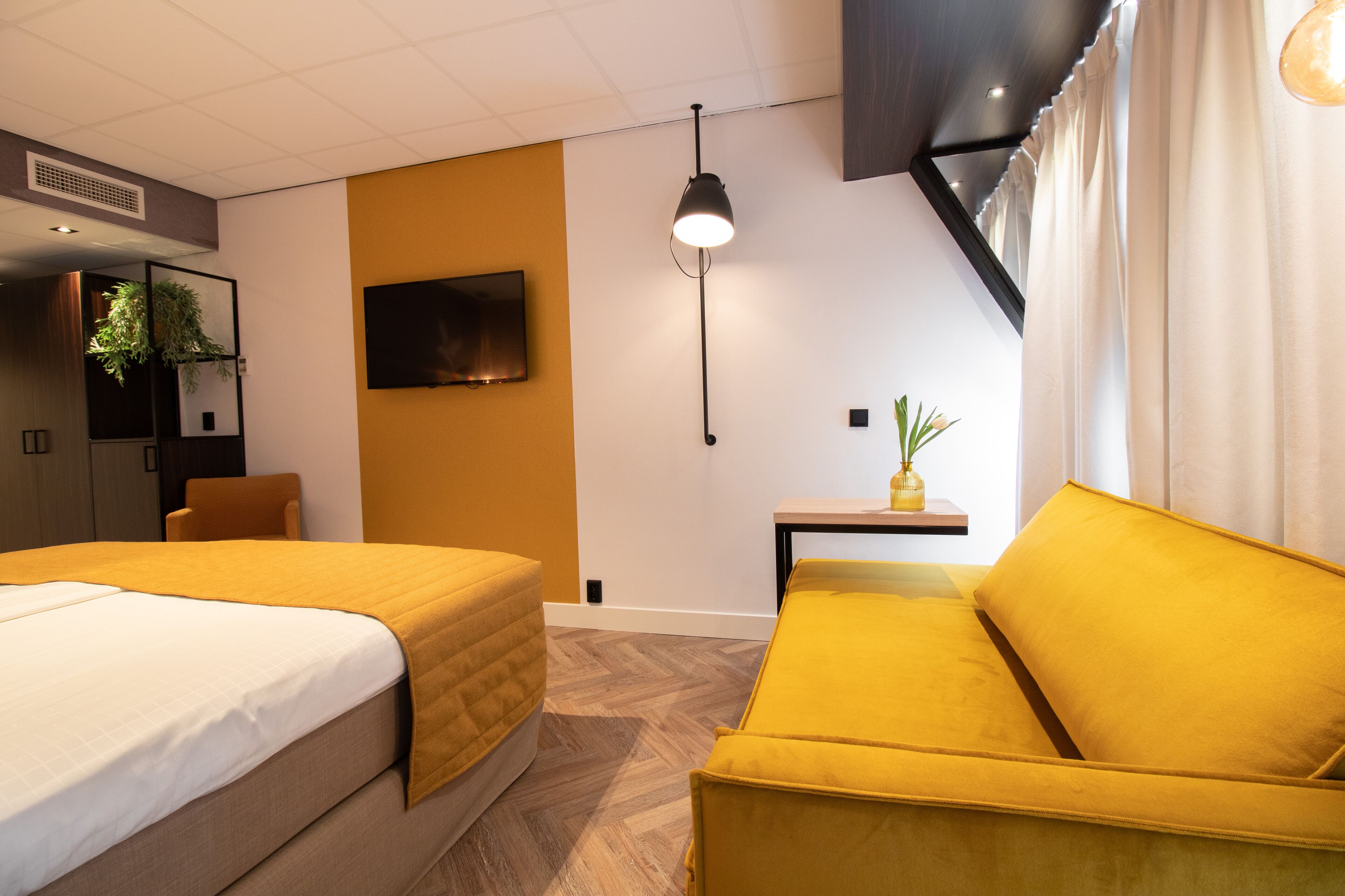 A hotel room with two beds, a sofa, a flat-screen TV, a small table with a plant, and a ceiling light.