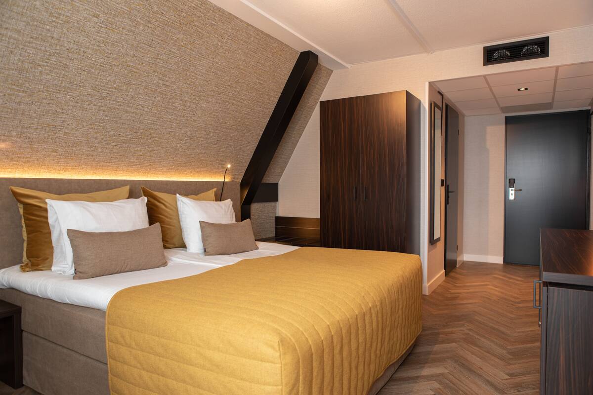 A modern hotel room with a large bed, a wooden wardrobe, and a door leading to another room.