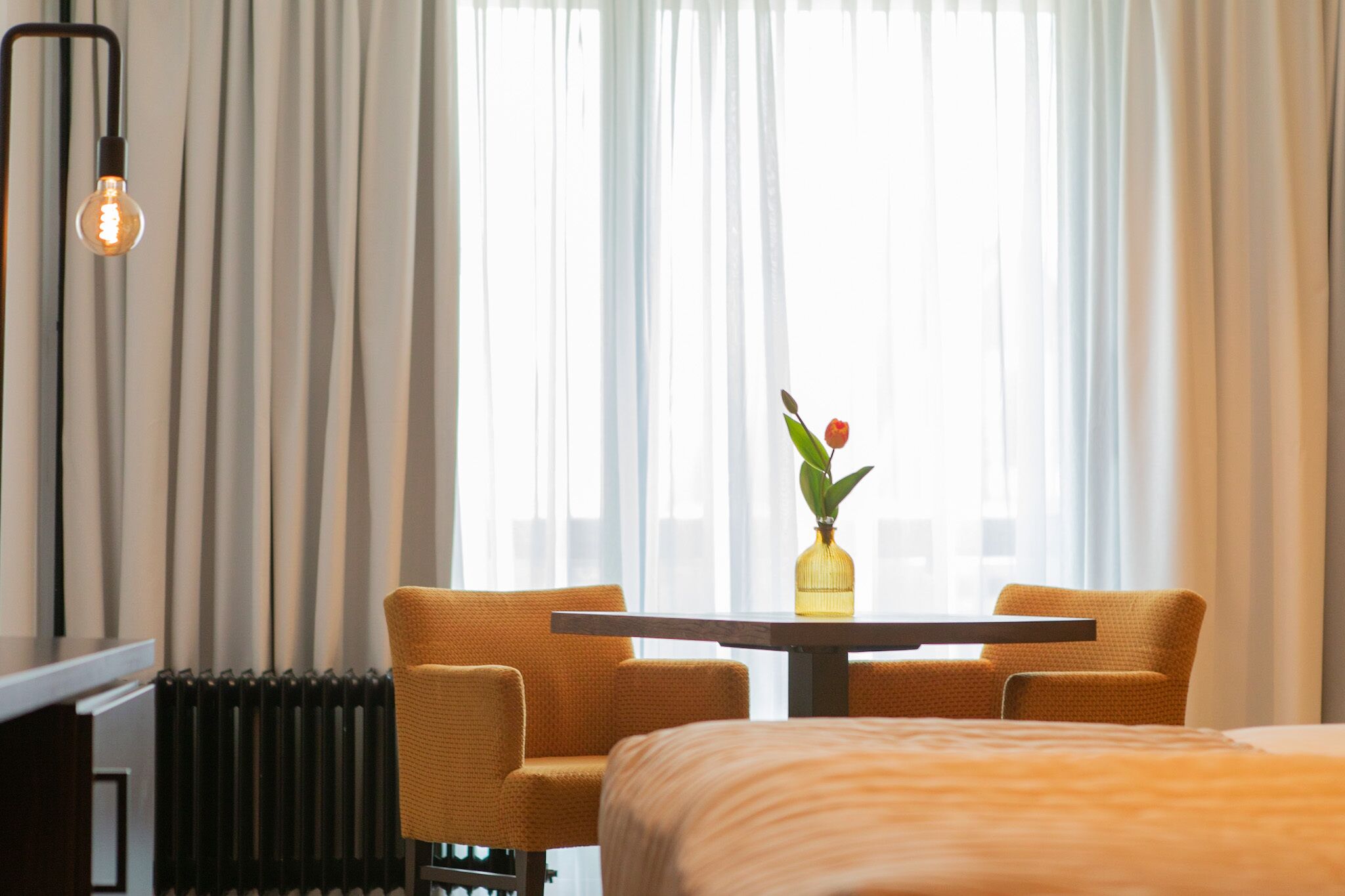 A hotel room with a bed, a table with two chairs, a vase with a flower, and a radiator.