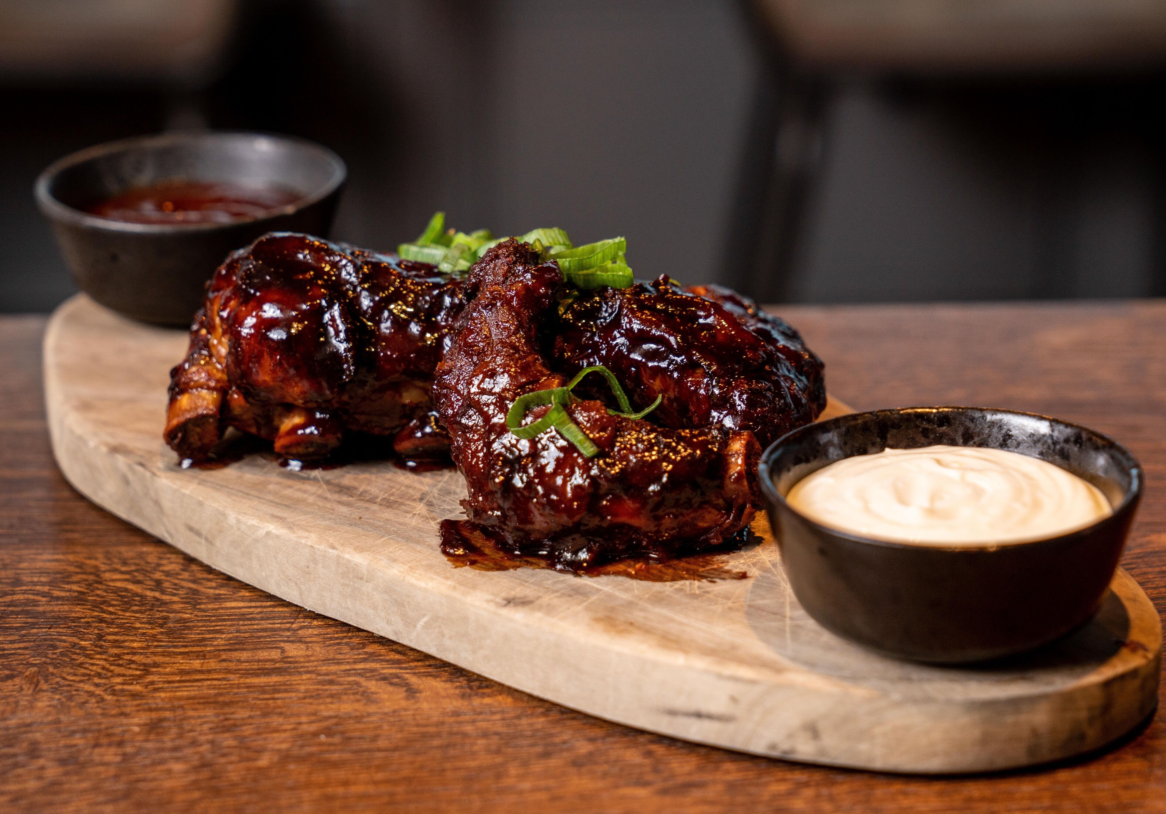 A wooden serving board with glazed meat and two small bowls of sauce.