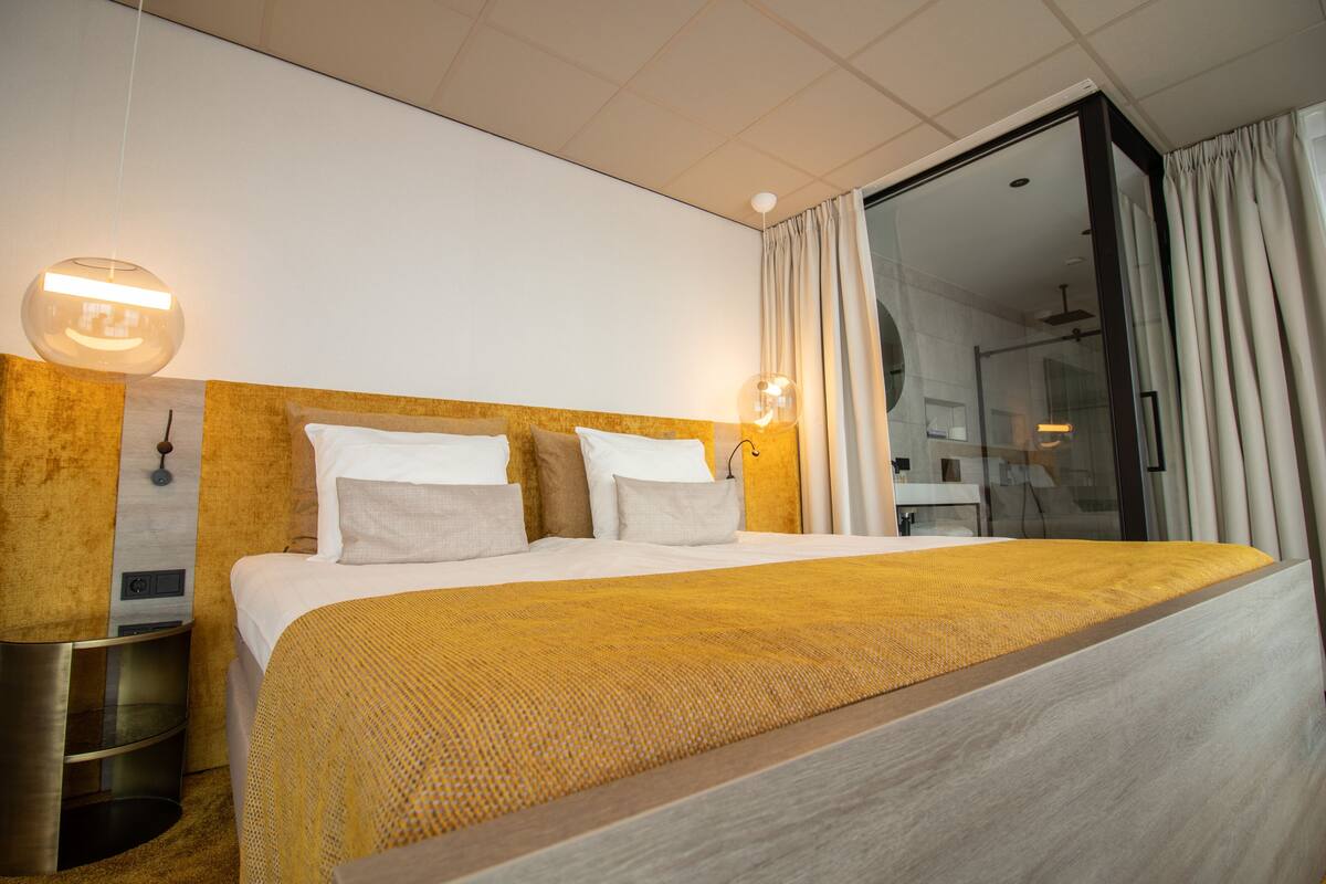 A modern hotel room with a large bed, a bedside table, a lamp, and a glass shower area.