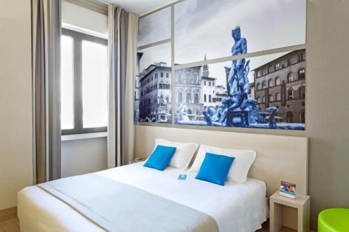 A hotel room with a bed, two pillows, a nightstand, and a large window.