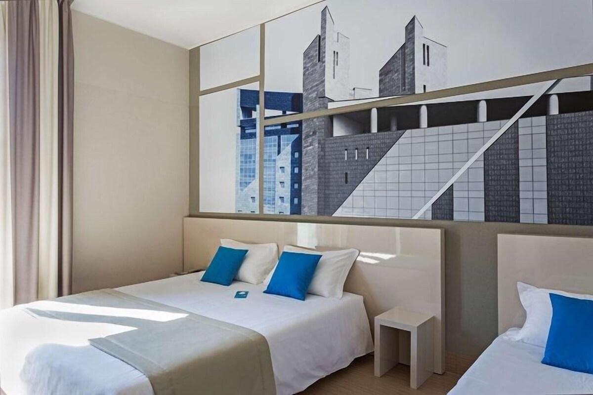 A hotel room with a large bed, a bedside table, and a view of a cityscape through a window.