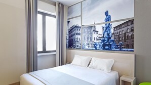 A hotel room with a large bed, a nightstand, a green chair, and a window with a view of a cityscape and a statue. - B&B Hotel Firenze Novoli (Florence)