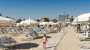 Uma praia com espreguiçadeiras e guarda-sóis, céu claro e edifícios ao fundo.