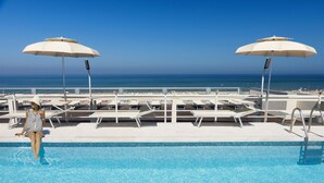 Uma área de piscina com espreguiçadeiras e guarda-sóis, vista direta para a praia e uma piscina com borda em azulejo.