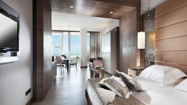 Quarto de hotel moderno com uma cama grande, televisão na parede, escrivaninha com cadeira e vista para a praia.
