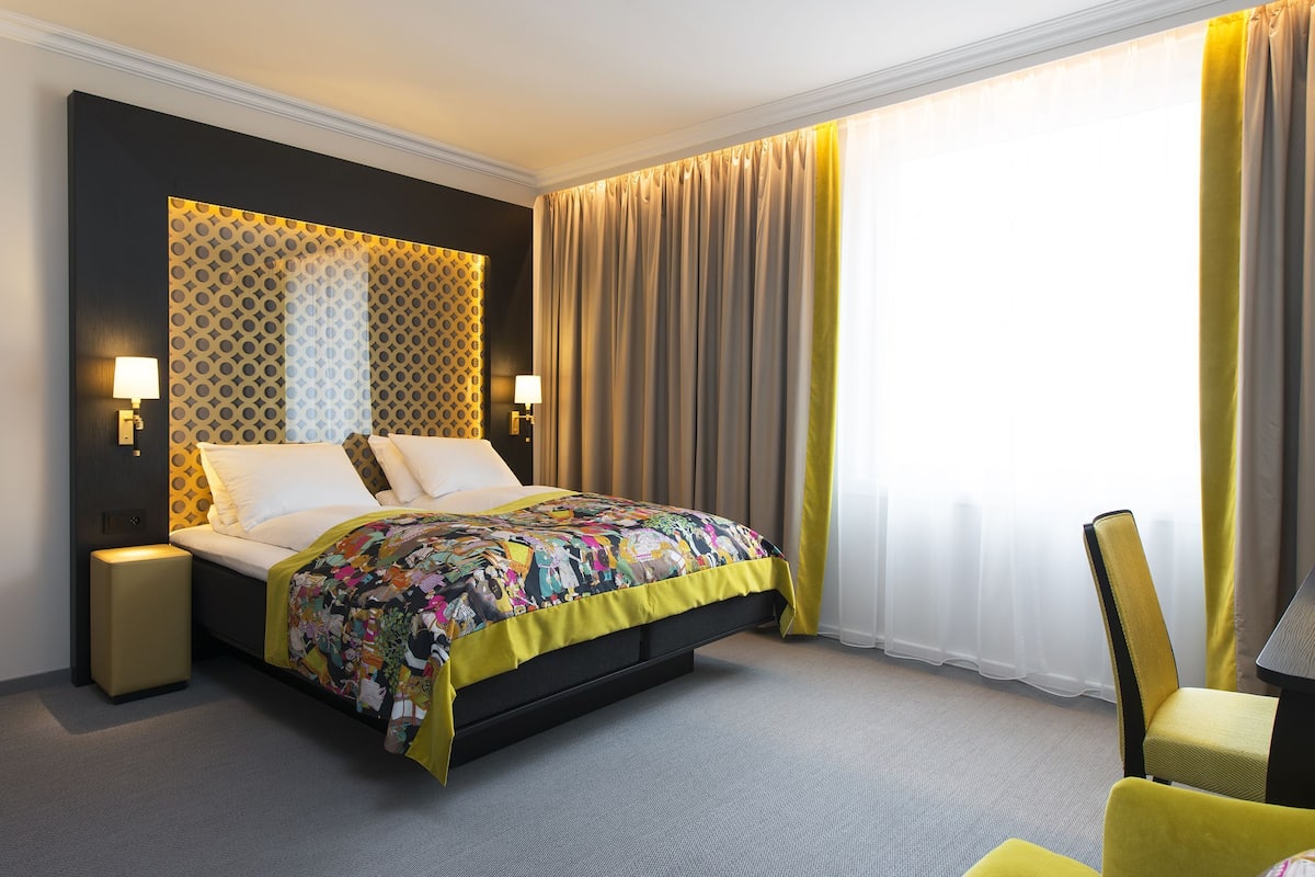 A hotel room with a bed, two bedside lamps, a chair, and a large window with curtains.