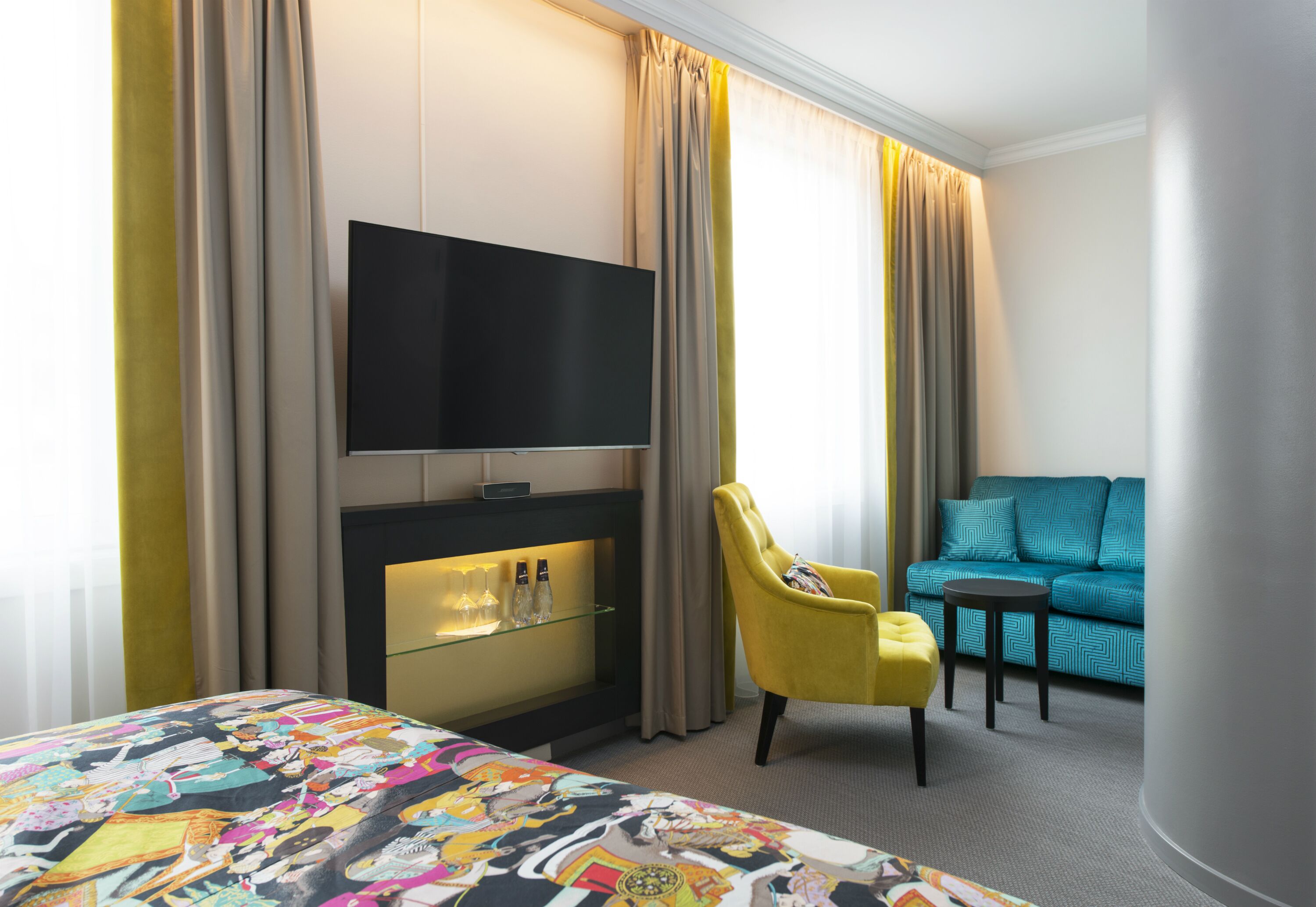 Habitación de hotel con cama, televisor montado en la pared, zona de estar con sofá y sillón, y mesa de vidrio con botellas.