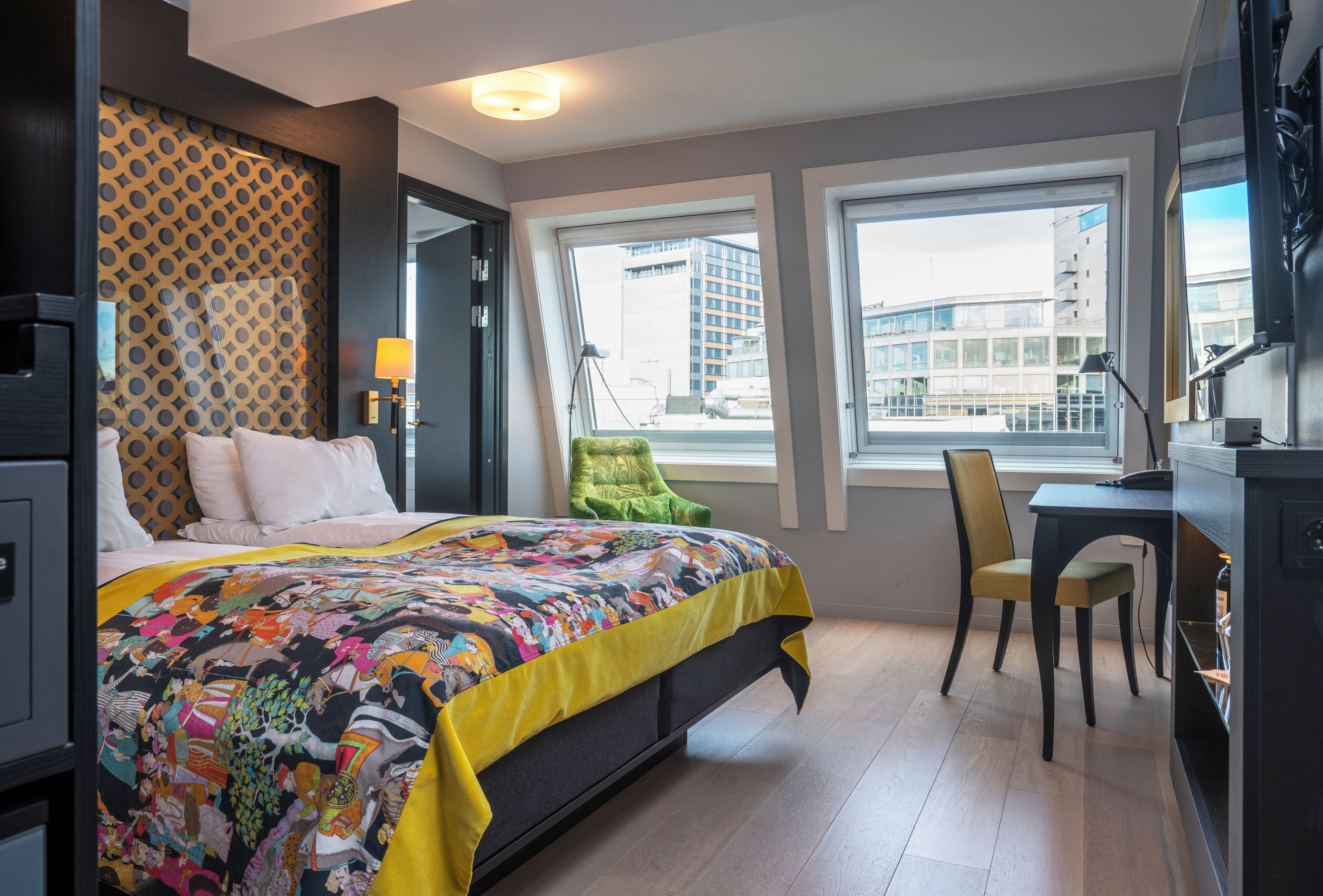 A hotel room with a bed, a desk, a chair, a window, and a view of buildings.