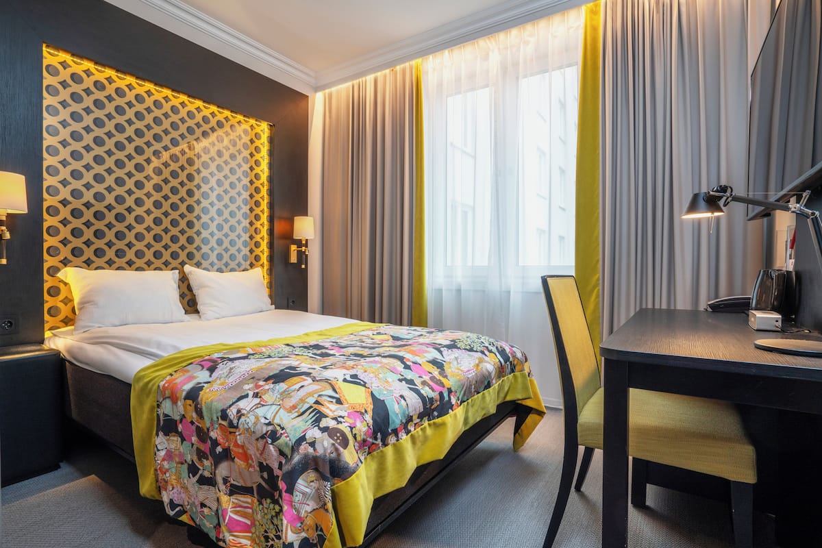 A hotel room with a bed, a desk, a chair, a lamp, and a patterned wall.