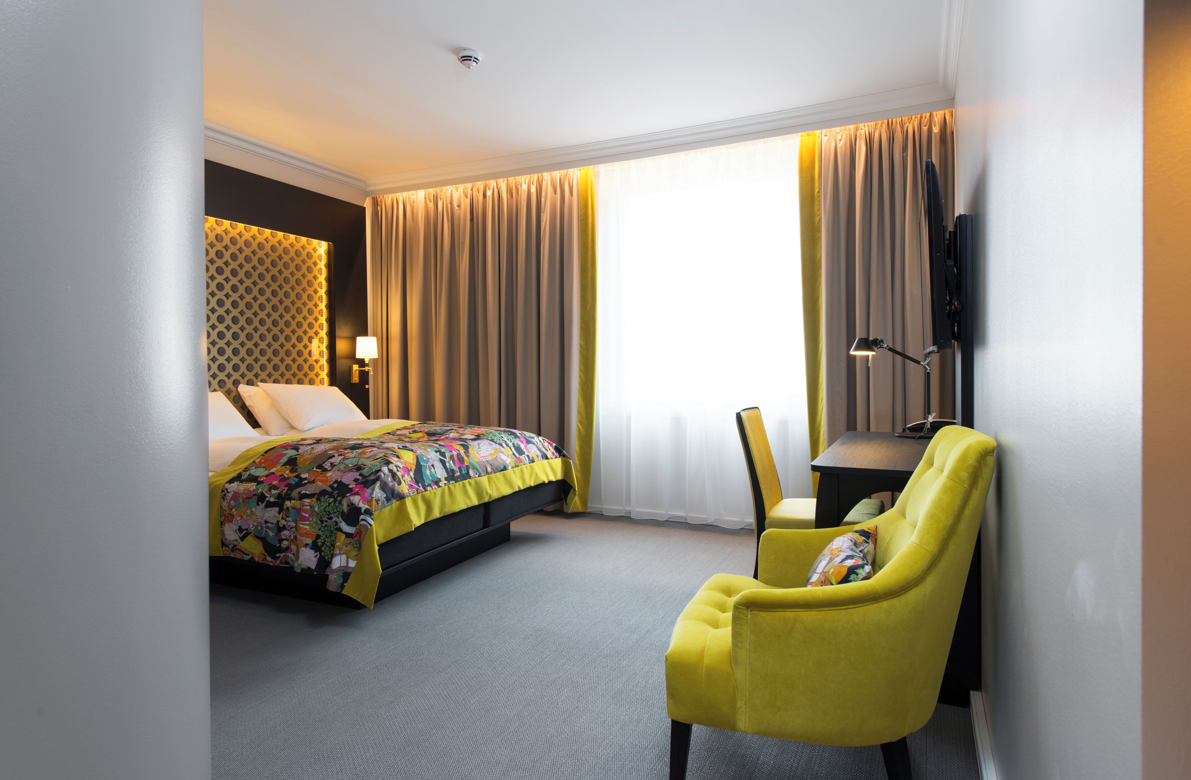 A hotel room with a bed, a yellow armchair, a desk, and a TV mounted on the wall.