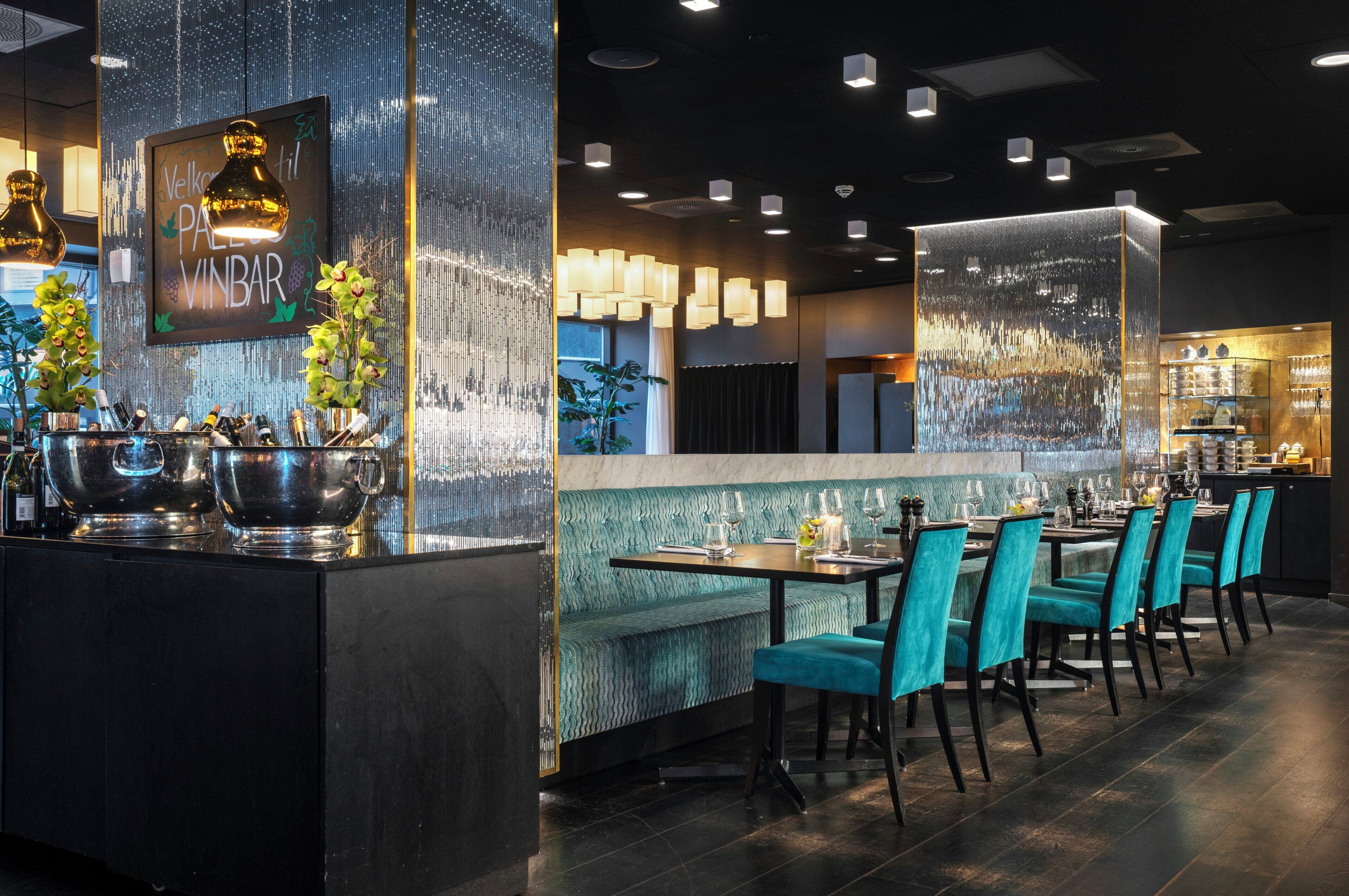 Restaurante com área de bar, assentos estofados em tons de verde-água e parede decorativa com um letreiro.