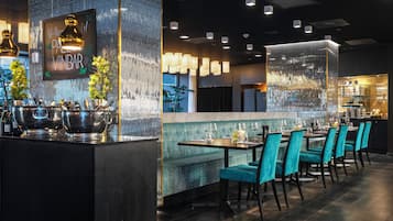 Restaurante com área de bar, assentos estofados em tons de verde-água e parede decorativa com um letreiro.
