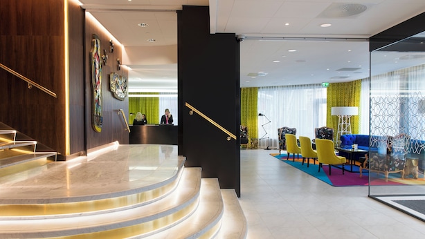 Eine Hotlellobby mit einer Rezeption, einer Treppe und einer Sitzecke mit Stühlen und einem Sofa.