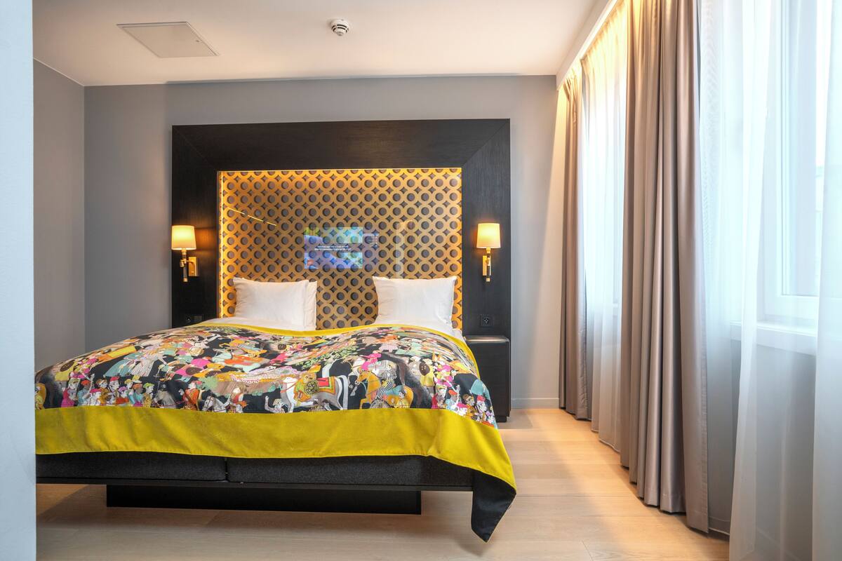 A hotel room with a large bed, a headboard with a geometric pattern, two wall-mounted lamps, and a window with curtains.