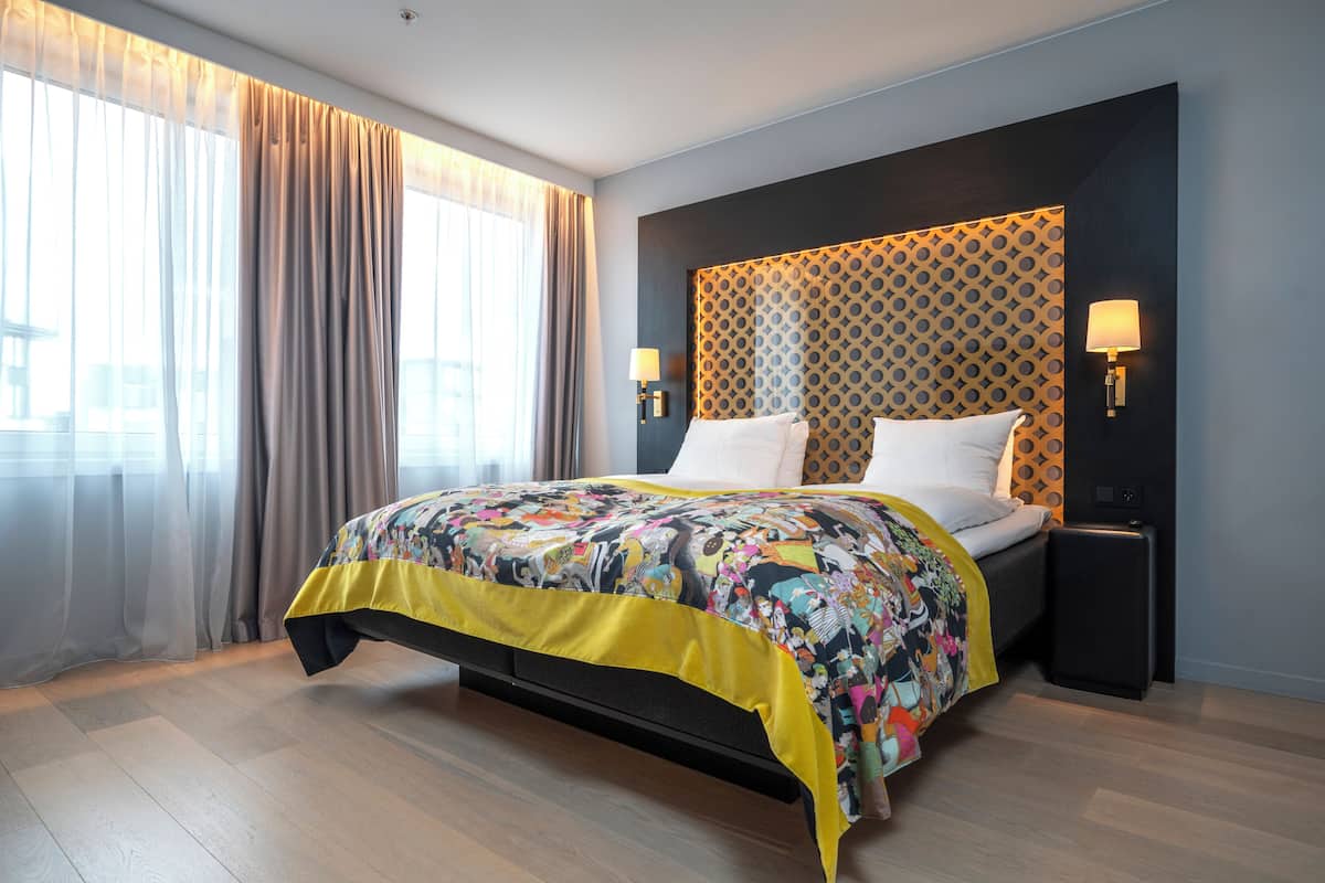 A modern hotel room with a large bed, a headboard with a geometric pattern, two bedside lamps, and a view through sheer curtains.