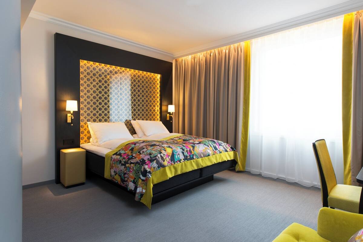 A hotel room with a bed, bedside lamps, a chair, and a window with curtains.