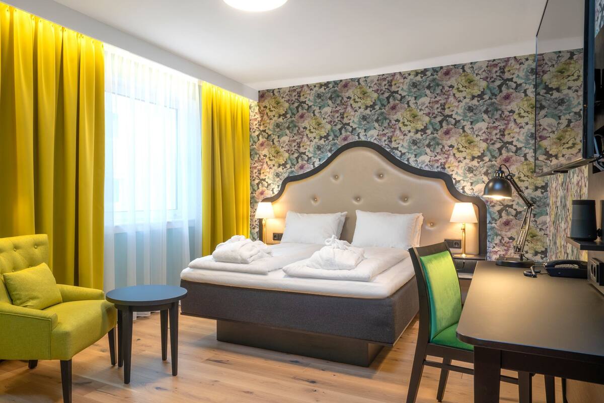 A hotel room with a bed, a green armchair, a small round table, and a dining table.
