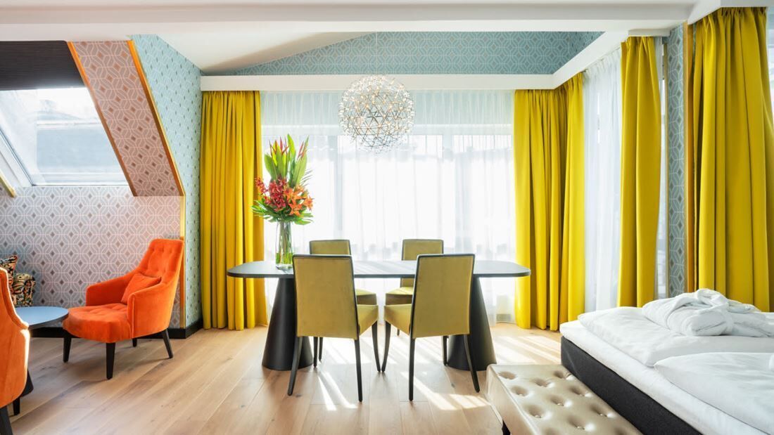 A hotel room with a bed, a dining table with chairs, a small table with a chair, a large window with curtains, and a decorative plant.