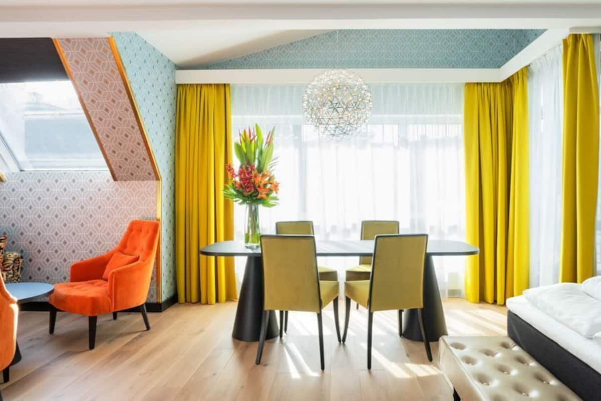 Una habitación de hotel con cama, una mesa de comedor con sillas, una mesita con una silla, un ventanal con cortinas y una planta decorativa.