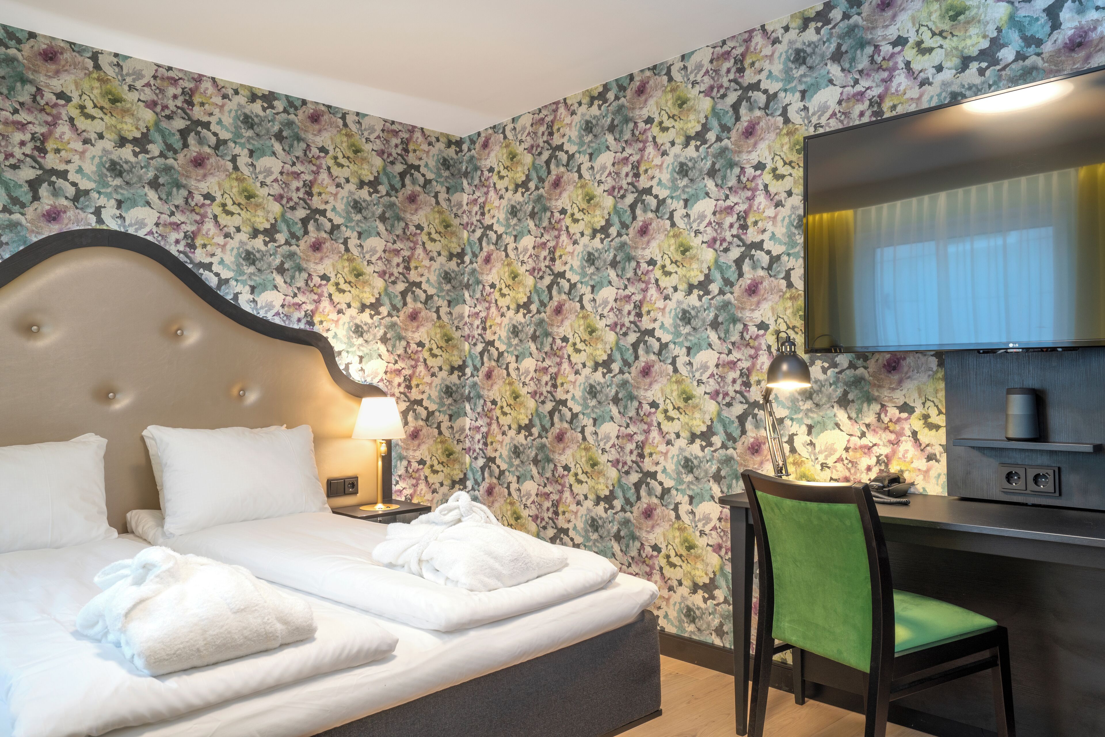 Habitación de hotel con cama, escritorio, silla, televisor y papel tapiz floral.