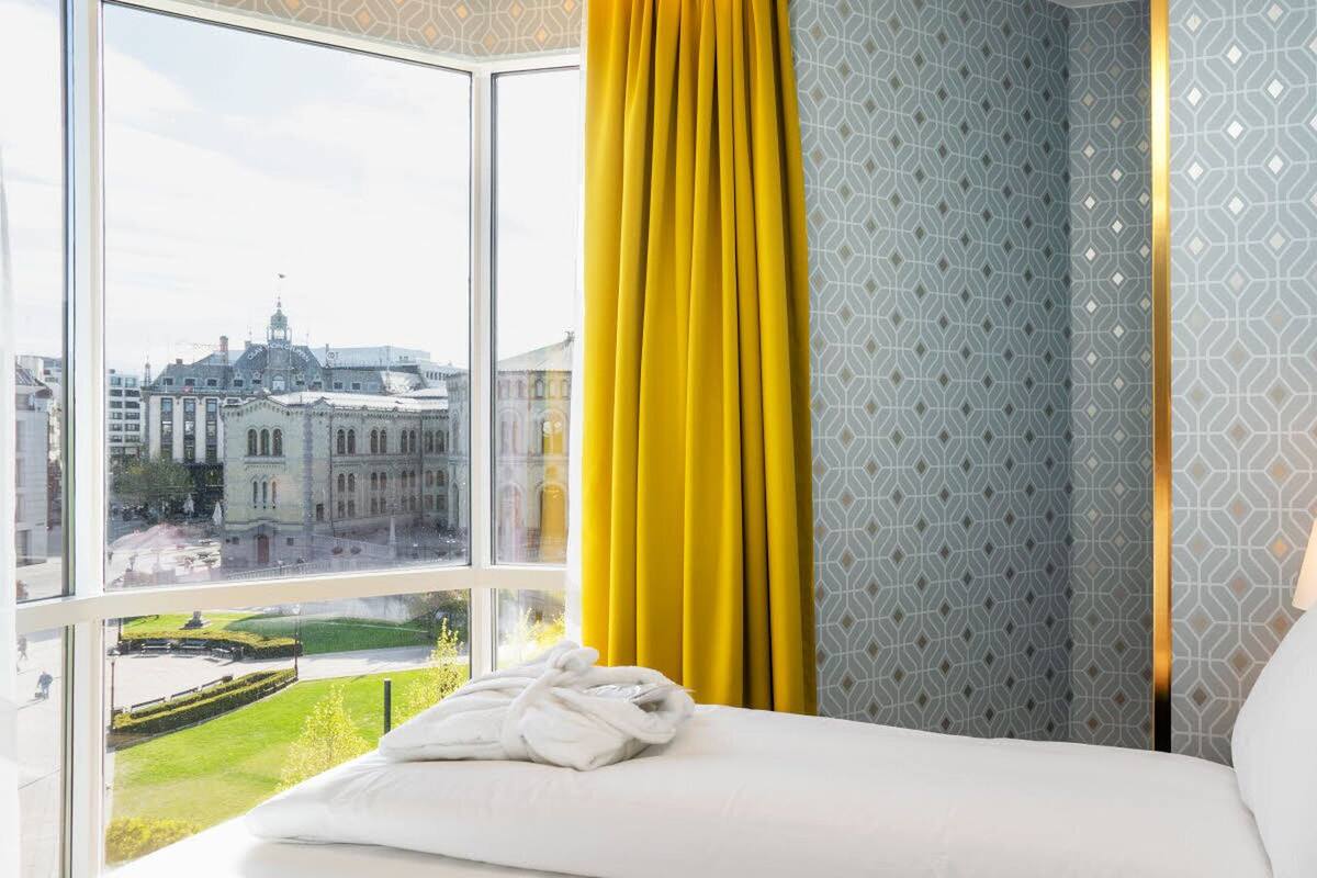 A hotel room with a large window, a bed with white linens, a yellow curtain, patterned wallpaper, and a view of a cityscape.