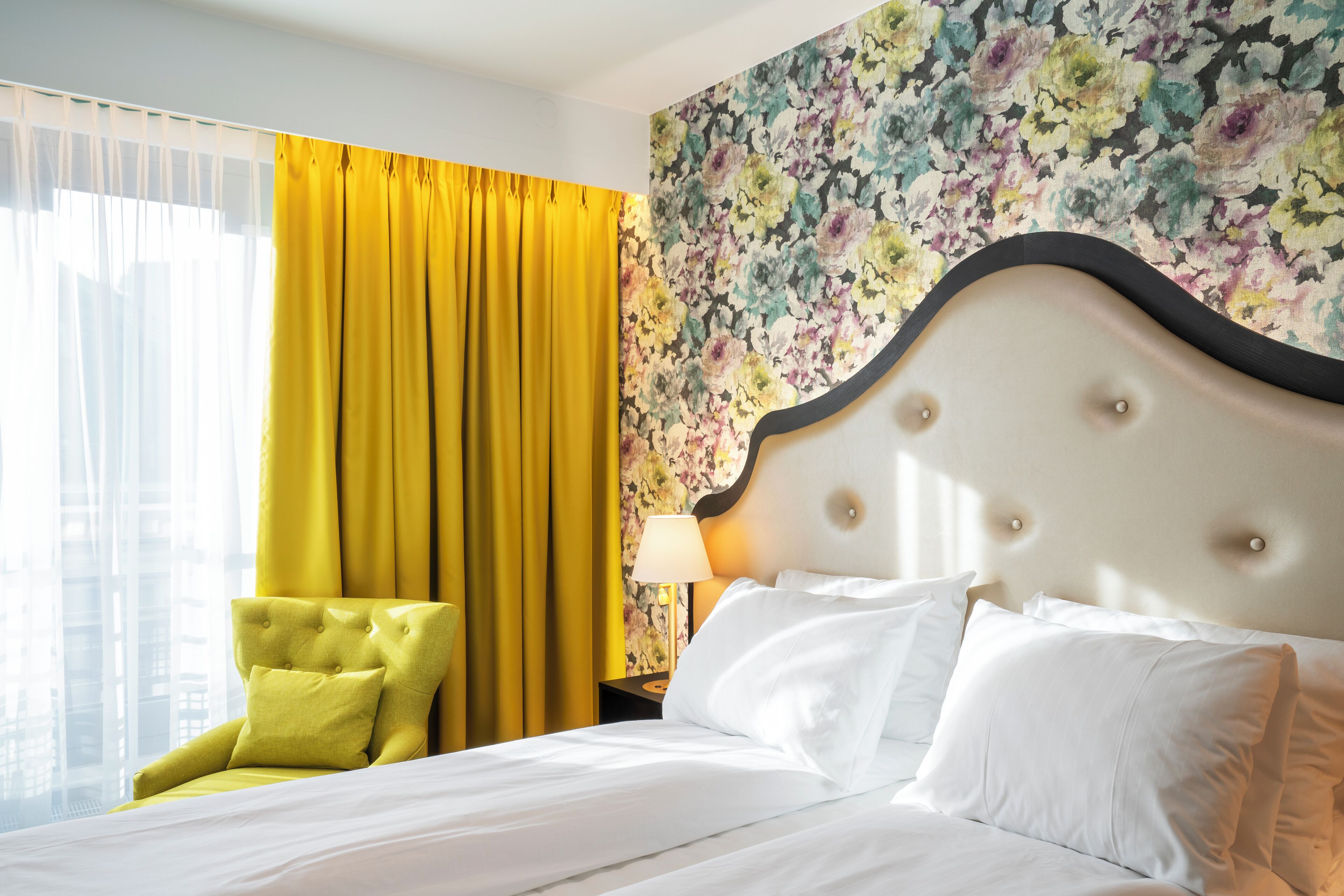 Une chambre d’hôtel avec un lit, une chaise jaune, un papier peint à motifs floraux et une fenêtre avec des rideaux jaunes.