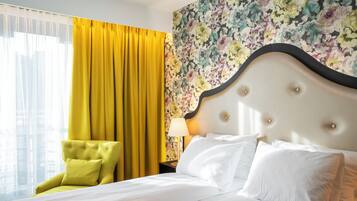Une chambre d’hôtel avec un lit, une chaise jaune, un papier peint à motifs floraux et une fenêtre avec des rideaux jaunes.