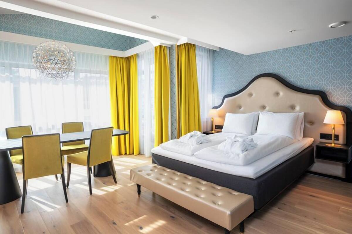A modern hotel room with a large bed, a bench, a dining table with chairs, and a chandelier.